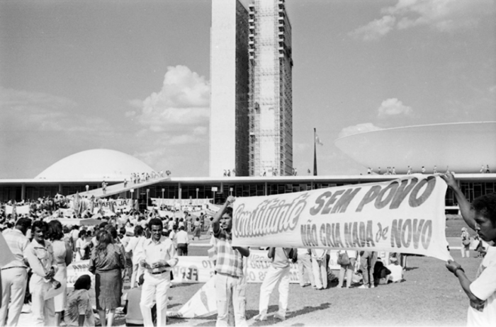 Foto em preto e branco. Diversas pessoas no Palácio do Planalto, em Brasília, subindo a rampa. No gramado, em primeiro plano, algumas pessoas seguram faixas nas quais é possível ler em uma delas: “Constituinte, sem povo não cria nada de novo.