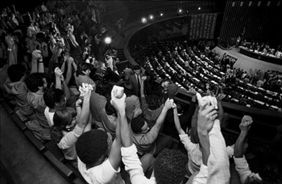 Foto em preto e branco pessoas na plateia de mãos dadas erguidas para o alto na sessão de votação do texto constitucional, que teve lugar no Plenário da Câmara dos Deputados.