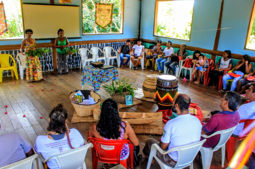 Foto com várias pessoas estão sentadas em cadeiras dispostas em semicírculo, numa sala grande, tendo ao centro, no chão, cestos, tambores e plantas.