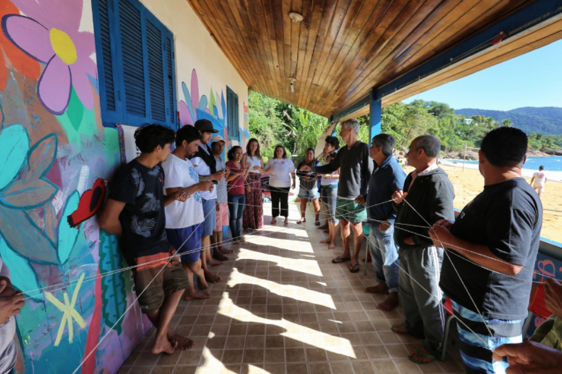 Foto de pessoas que participam de uma dinâmica. Elas estão de pé, umas de frente para as outras,  interligadas por um barbante, que foi esticado para que cada pessoa pudesse segurar numa ponta, formando uma rede.