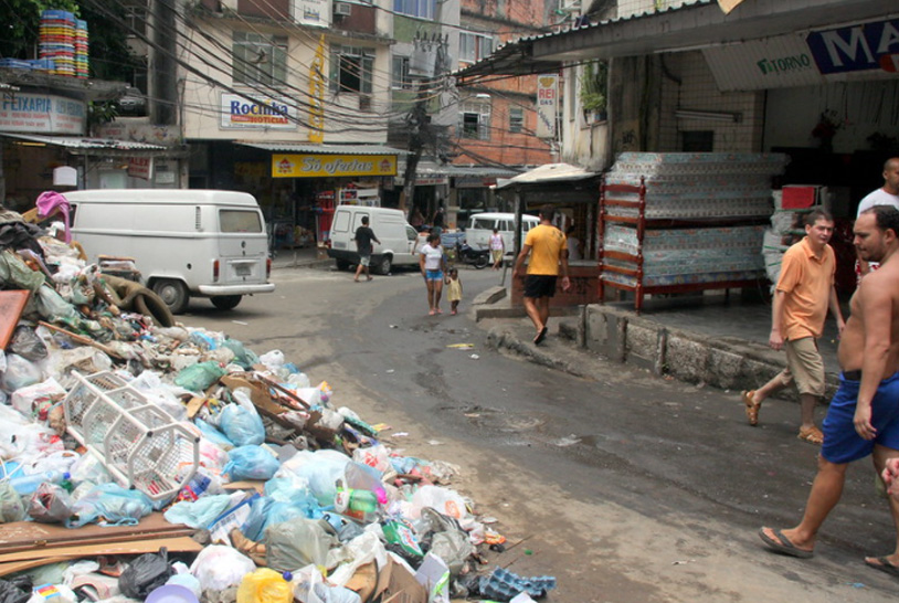 Foto de um homem caminhando numa rua e observando uma pilha de lixo a céu aberto.
