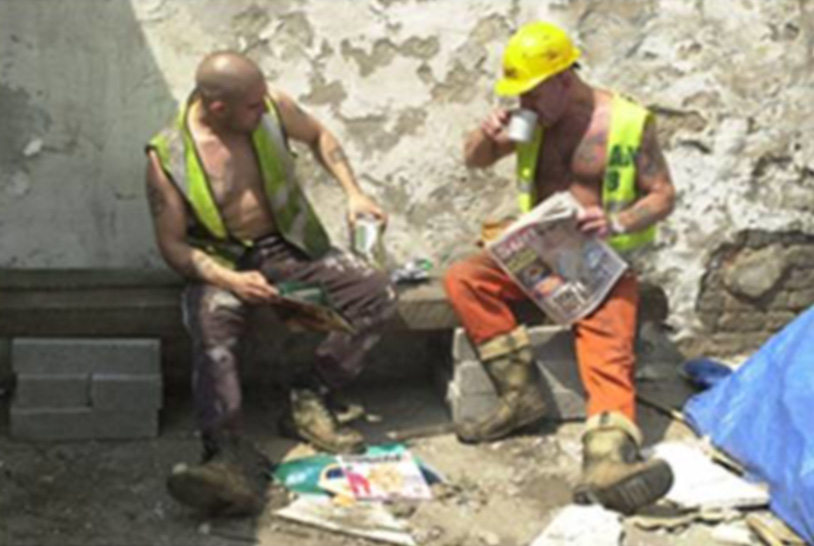 Um banco com dois trabalhadores sentados vestindo coletes, calças, botas e capacete, sujos do trabalho, fazendo uma pausa para um café.