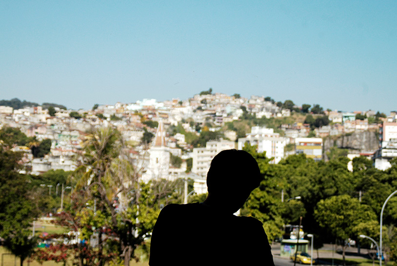 Sombra de um homem que está olhando para um morro com diversas construções que remetem a uma favela.